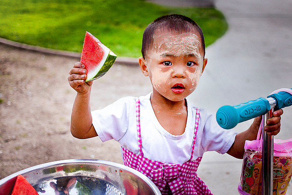 A little girl who is a Burmese refugee holds a slice of watermelon next to her scooter.